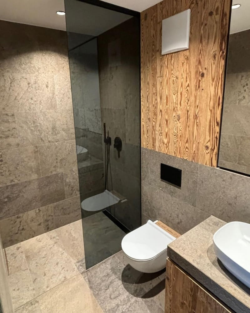 Modernes Badezimmer mit grauen Steinfliesen an den Wänden und einem wandmontierten WC. Links befindet sich eine begehbare Dusche mit einer gläsernen Trennwand. Die rechte Seite des Raumes ist mit einer Holzverkleidung ausgestattet, die dem Raum eine warme und natürliche Atmosphäre verleiht