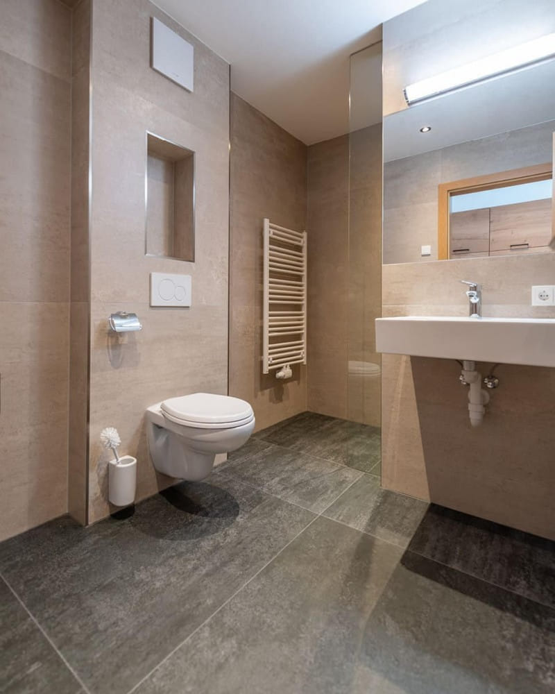 Modernes Badezimmer mit hellen Wandfliesen und dunklem Steinboden. Rechts ist ein großes Waschbecken mit Spiegel und darüber eine lange Leuchte angebracht. Das Design ist schlicht und funktional, mit klaren Linien und einer neutralen Farbpalette.