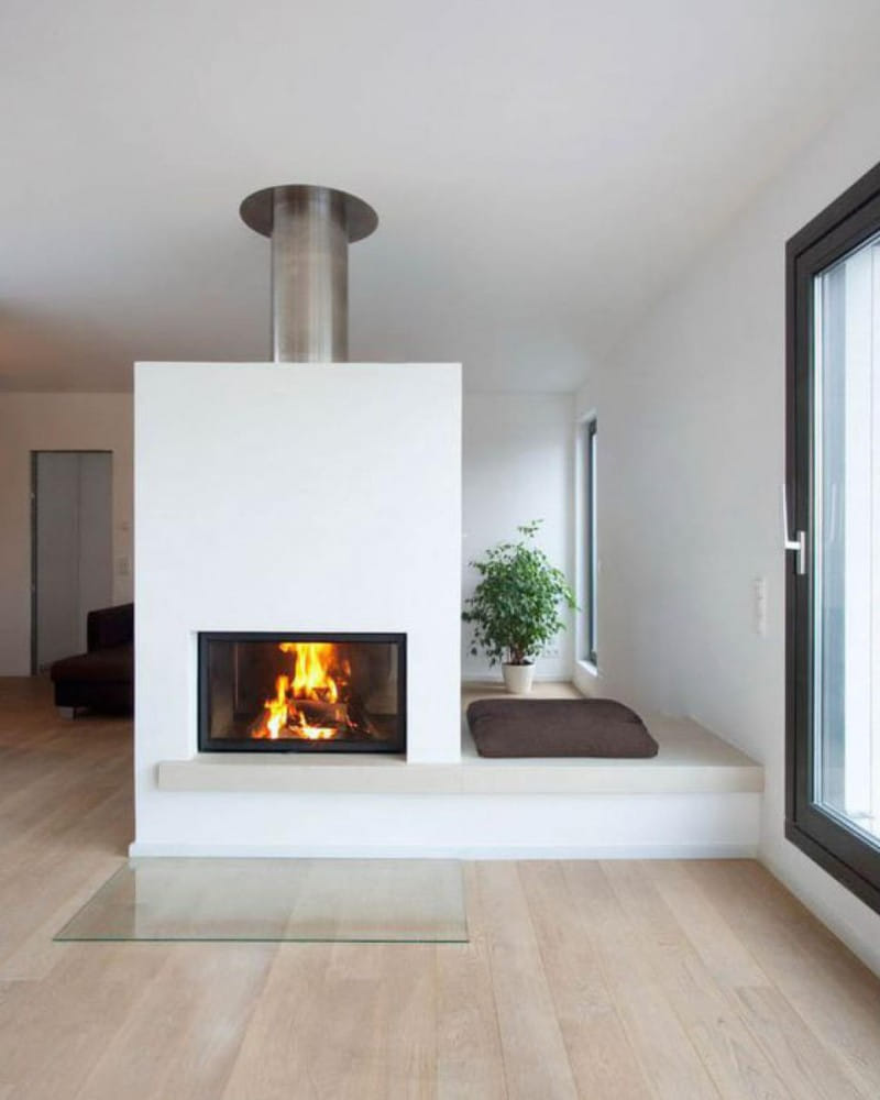 Minimalistisch gestalteter Wohnbereich mit einem modernen Kamin in der Mitte des Raums. Der Kamin hat ein schlichtes, weißes Design mit einem Metallabzug und ein brennendes Feuer im Inneren. Rechts vom Kamin befindet sich eine Sitzbank mit einem Kissen und einer Zimmerpflanze als Dekoration.