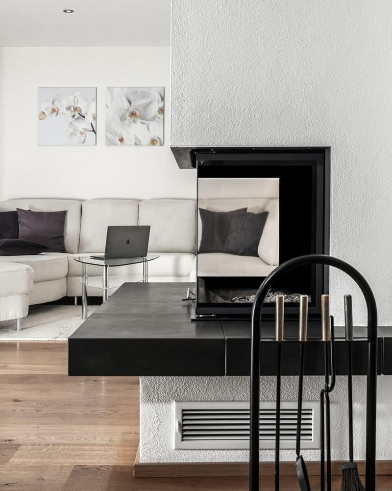 Modernes Wohnzimmer mit einem eckigen Kamin in schwarzem Design. Im Vordergrund befinden sich Kaminwerkzeuge, und im Hintergrund ist eine helle, L-förmige Sofalandschaft mit dunkelgrauen Kissen zu sehen.