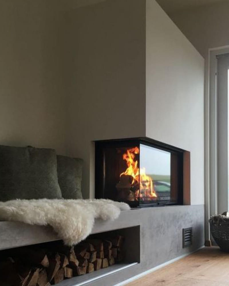 Gemütlicher Wohnbereich mit modernem, eckigem Kamin, in dem ein Feuer brennt. Rechts neben dem Kamin befindet sich ein Fenster, das natürliches Licht hereinlässt. Vor dem Kamin liegt ein Sitzbereich mit grauen Kissen und einem weichen, weißen Fell. Unter der Sitzbank sind gestapelte Holzscheite als dekoratives Element aufbewahrt.