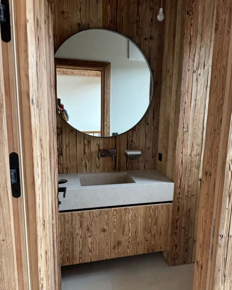 Badezimmer mit einer Wandverkleidung aus hellem Holz. Über dem rechteckigen Waschbecken aus Naturstein hängt ein großer, runder Spiegel, der den Raum optisch vergrößert. Die Einrichtung kombiniert natürliche Materialien mit modernen Elementen wie einem minimalistischen Waschbecken und einer in die Wand integrierten Armatur.